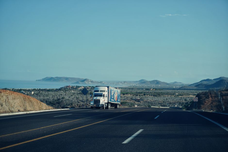 Infraestrutura Logística — A semi-truck travels on a scenic highway with mountains in the background under a clear sky.